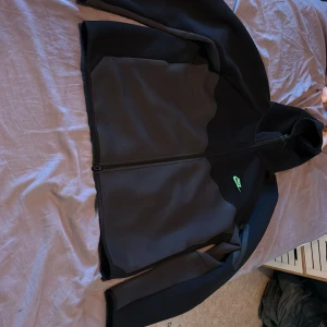 Svart Nike Tech fleece med huva L - Nike tech fleece i svart med diskret grå detalj och neongrön logga på bröstet. Jackan har hel dragkedja, huva och praktiska fickor med dragkedja på ärmen. Snygg och sportig look, perfekt för dig som gillar streetwear.