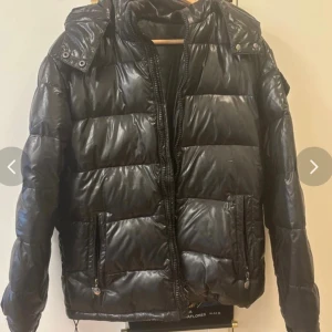 Svart pufferjacka från Fashion M - Säljer en svart, glansig pufferjacka från Fashion M med huva och dragkedja framtill. Jackan har två fickor med dragkedja och en patch med logga på ärmen. Perfekt för kalla dagar och har en riktigt snygg streetstil.