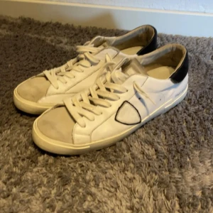 Vita sneakers från Philippe Model - Snygga vita sneakers från Philippe Model med svart häl och klassisk snörning. Skorna har en rund tå, platt sula och är tillverkade i skinn. Diskret logga på sidan och neutrala färger gör dem lätta att matcha med olika outfits.(jag går inte under 1600kr köpta för 2500)