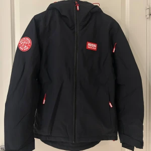 Svart skidjacka från Huski Wear - Svart skidjacka från Huski Wear med huva och röda detaljer. Använd i två säsonger och är fortfarande i bra skick. Finns flera fickor för liftkort. Pris går att diskuteras.