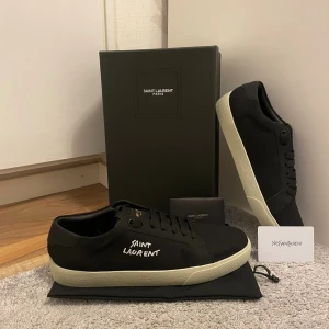 Svarta sneakers från Saint Laurent - Svarta sneakers från Saint Laurent med vit logga broderad på sidan och vit platt sula. Skorna har klassisk låg profil, snörning och är tillverkade i canvas med detaljer i skinn. Perfekta för dig som gillar stilren och lyxig streetstyle.