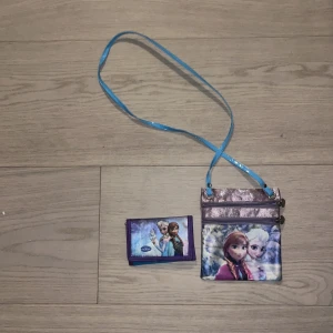 Frozen barnaxelväska och plånbok - Säljer en söt axelväska med motiv från Frozen och matchande plånbok. Väskan har blå axelrem, två dragkedjor och glittrig lila framsida. Plånboken är lila med snöflingor och Frozen-tryck. Perfekt för barn som älskar Disney och vill ha med det viktigaste.