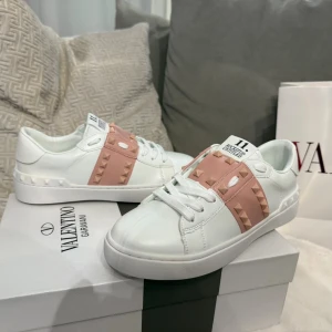 Valentino Rockstud vita sneakers - Snygga vita Valentino Garavani Rockstud sneakers med ljusrosa band och ikoniska nitar. Skorna har rund tå, platt sula och snörning. Tillverkade i skinn med coola detaljer och en modern vibe. Perfekta för dig som vill sticka ut med exklusiv streetstyle.