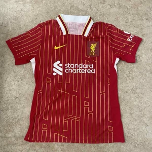 Liverpool FC röd matchtröja Nike - Liverpool FCs officiella matchtröja i rött med gula ränder, klubbmärke och Nike-logga på bröstet. Tröjan har vit och gul detalj vid kragen och sidorna samt sponsortryck framtill. Tillverkad i lätt och ventilerande material.
