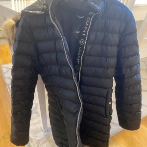 Svart lång dunjacka från GANT i storlek 170 - Säljer en svart, lång dunjacka från GANT med snyggt quiltad design och huva med fuskpäls. Jackan har dragkedja framtill, tryckknappar vid fickorna och GANT-logga på bröstet. Perfekt för kalla dagar och riktigt stilren look.