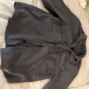 Grå overshirt från H&M i ullmix - Säljer en stilren grå overshirt från H&M i ullmix. Skjortan har två stora bröstfickor med lock, knäppning framtill och klassisk krage. Perfekt lager-på-lager-plagg med lång ärm och enkel, clean look.