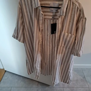 Randig kortärmad skjorta från 157 - Beige och vit randig skjorta från Lager  157 med korta ärmar, bröstficka och klassisk krage. Skjortan är tillverkad i ett luftigt linnematerial och har knappar framtill. Perfekt för en avslappnad och trendig look skjortan blev aldrig använd nypris 150.