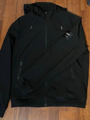 Svart jacka med dragkedja och huva - Svart hoodie från Jack and Jones med dragkedja framtill, två fickor med dragkedja och justerbar huva. Klassisk och stilren design med ribbade muddar och liten logotyp på ärmen. Perfekt för en avslappnad och sportig look. Vi kan diskutera pris.