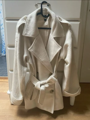 Beige kappa med bälte och stora slag - Stilren beige kappa med breda slag, dubbelknäppning och knytskärp i midjan. Kappan har axelklaffar och stora fickor, perfekt för en trendig look. Materialet är mjukt och ger en lyxig känsla. Passar perfekt till höst, vår och vinter. Priset kan diskuteras. 