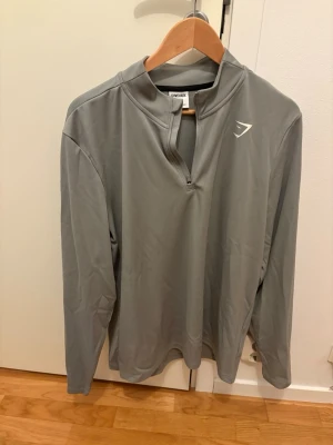 Grå långärmad träningströja Gymshark - Snygg grå långärmad träningströja från Gymshark med halv dragkedja och hög krage. Tillverkad i ett lätt och stretchigt funktionsmaterial som passar perfekt till gymmet eller löprundan. Diskret logga på bröstet.