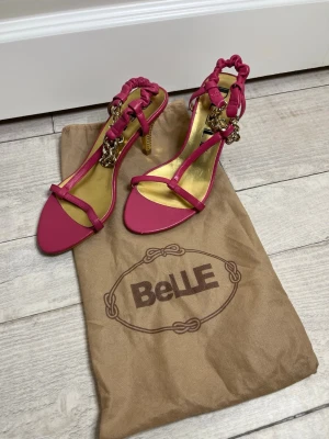 Rosa pumps med gulddetaljer från BeLLE - Snygga rosa pumps i storlek 36 men passar också 37. Från BeLLE i äkta fårskinn med smala remmar och guldfärgad innersula. Klackarna är dekorerade med guldfärgade kedjor och detaljer, och remmarna runt ankeln är vadderade för extra stil. Perfekta för dig som gillar att sticka ut. 