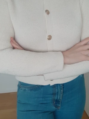 Krämvit stickad kofta med fickor - Säljer en krämvit stickad kofta med snygga guldknappar framtill och på de två små fickorna. Koftan har lång ärm och rak passform.