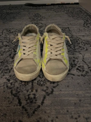 Philippe Model sneakers med gula detaljer - Snygga sneakers från Philippe Model i beige med gula detaljer och vita snören. Skorna har lågt skaft och klassisk siluett. Tillverkade i en mix av läder och textil med gummisula. Perfekta för dig som gillar streetstyle och vill sticka ut lite extra.