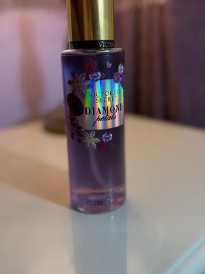 Victoria's Secret Diamond Petals parfym - Säljer en Victoria's Secret Diamond Petals parfym i en snygg lila flaska med blommiga detaljer och guldig kork. Flaskan rymmer 250 ml och har en holografisk etikett som ger en lyxig känsla. Perfekt för dig som gillar fräscha och blommiga dofter.