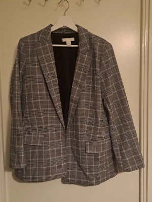 Grå rutig kavaj från H&M - Grå kavaj med rutigt mönster från H&M. Tyget är sånt gammaldagsstil så det kan se ut som att det är lite nopprigt, men det är en del i designen. Kavajen har två framfickor med lock, smal krage och helfodrad insida. 🩷