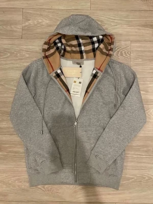 Grå Burberry Zip Up - Snygg grå hoodie från Burberry med klassiskt rutigt foder i huvan. Tröjan har dragkedja framtill, dragsko i huvan och ribbade muddar vid ärmslut och nederkant. Perfekt för en stilren och trendig look. Materialet är mjukt och bekvämt.