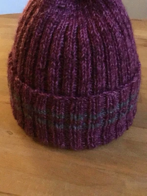 Lila stickad mössa med randig kant - Säljer en ribbstickad lila mössa med uppvikt kant och diskreta ränder i gråt och lila. Mössan är gjord i ett mjukt, garnigt material och har en klassisk form som sitter skönt på huvudet. Perfekt för kalla dagar och snygg till jackan.