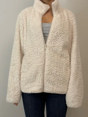 Krämvit teddyjacka med dragkedja - Mysig teddyjacka i krämvit färg med hög krage och dragkedja framtill. Jackan har en fluffig och mjuk textur som ger en avslappnad och trendig look. Perfekt för kyliga dagar och enkel att matcha med jeans eller leggings.