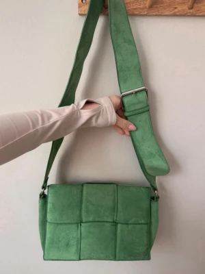 Grön axelväska i flätad mocka-look - Trendig grön axelväska med bred justerbar rem och flätad design i mockaimitation. Väskan har en rektangulär form och stängs med lock. Perfekt för dig som vill ha en statement piece till din outfit.
