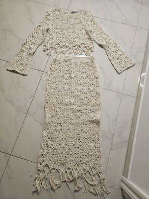 Beige virkat set med fransar - Snyggt tvådelat set i beige virkat material med långärmad topp och lång kjol. Toppen har vågig kant och kjolen har fransar nertill. Perfekt för en bohemisk look. Setet är handgjort i bomullsliknande garn och har ett unikt mönster.