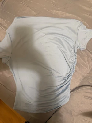 Ljusblå topp med rynkdetalj - Säljer en ljusblå topp med korta ärmar och snygg rynkdetalj på sidan som ger en cool twist till outfiten. Perfekt att matcha med jeans eller kjol för en fräsch look.