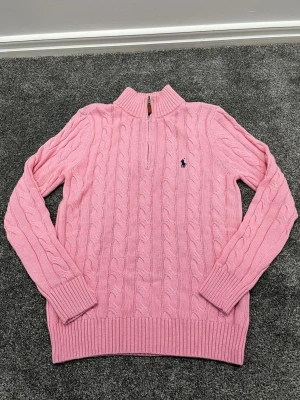 Rosa kabelstickad tröja Polo Ralph Lauren - Kabelstickad rosa tröja från Polo Ralph Lauren med halv dragkedja och ribbade muddar. Tröjan har en liten mörkblå logga broderad på bröstet och är tillverkad i mjukt material. Perfekt för en avslappnad men ändå stilren look.