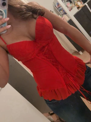 Röd korsett top - Säljer en snygg röd korsett top. Modellen har vadderade kupor och figurnära passform som framhäver midjan. Perfekt för dig som vill ha en statement piece i garderoben.