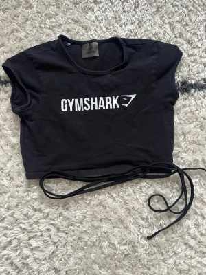 Svart croppad topp från Gymshark - Svart croppad topp från Gymshark med vit logga framtill. Toppen har korta ärmar och långa snören för knytning i midjan. Perfekt för träning eller gymmet. Tillverkad i mjuk bomull som känns skön mot huden.
