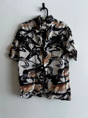 skjorta - Kortärmad skjorta med abstrakt svart, brun och vit mönsterdesign. Skjortan har klassisk krage och knäppning framtill. Perfekt för dig som gillar en unik och konstnärlig stil. Materialet ser ut att vara lätt och luftigt, perfekt för varmare dagar.