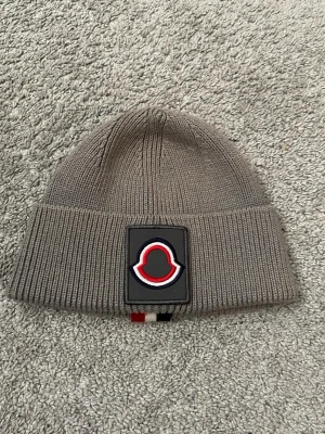Grå Moncler mössa - Snygg grå stickad mössa från Moncler med bred uppvikt kant och stor svart patch med Moncler-logga i rött och vitt framtill. Baksidan har en rand i svart, vitt och rött. Perfekt för dig som gillar streetstyle och exklusiva märken.