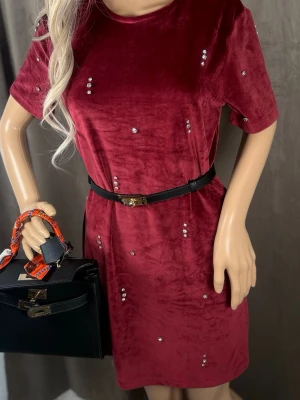 Vinröd sammetsklänning med pärlor - Vinröd kortklänning i mjuk sammet med pärldetaljer och rund hals. Klänningen har korta ärmar och ett svart bälte i midjan som ger en snygg siluett. Perfekt för dig som vill ha en unik och trendig look.