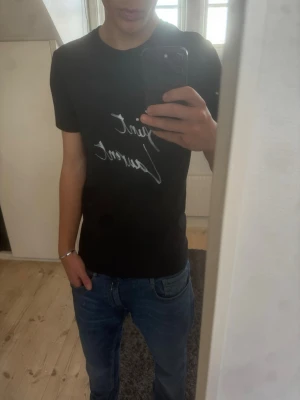 Svart Saint Laurent t-shirt - Säljer en svart t-shirt från Saint Laurent med vit logga i handskriven stil på bröstet. Klassisk passform och rund halsringning. Perfekt till jeans eller shorts för en clean och stilren look.