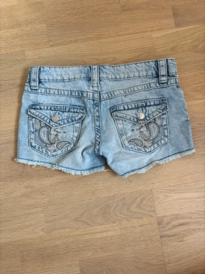 Ljusblå jeansshorts med broderi - Säljer ett par ljusblå jeansshorts från Gina Tricot med slitna detaljer och rå kant. Shortsen har broderade bakfickor med silverfärgade knappar och bälteshällor. Perfekta för sommaren och ger en avslappnad look.