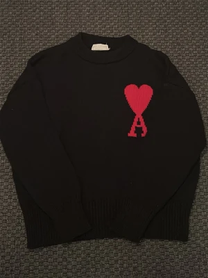 Svart stickad tröja från Ami Paris, pris kan diskuteras och fler bilder kan skickas  - Svart stickad tröja från Ami Paris med röd hjärtlogga och bokstaven A på bröstet. Tröjan har rund halsringning, långa ärmar och ribbstickade muddar. Perfekt för dig som gillar stilrena plagg med en twist.