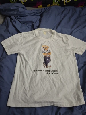 Vit Polo Bear t-shirt från Ralph Lauren - Vit t-shirt från Ralph Lauren med ikoniskt Polo Bear-motiv på bröstet. Modellen är Custom Slim Fit och har rund halsringning. Trycket visar en björn i randig tröja och blå byxor. Perfekt för dig som gillar klassisk och lekfull stil.