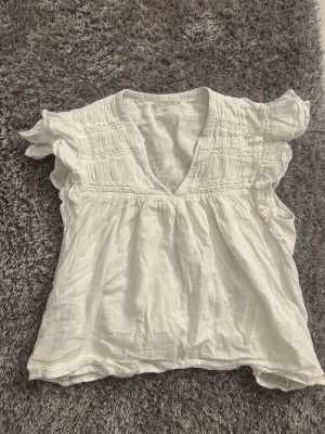 Vit blus med volangärmar och v-ringning - Söt vit blus med v-ringning och volangärmar. Blusen har smockdetaljer upptill och är tillverkad i ett lätt, luftigt material som känns perfekt för varmare dagar. Croppad passform som ger en avslappnad och trendig look.