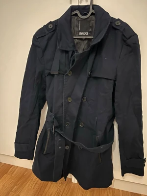 Mörkblå trenchcoat från Smog - Stilren mörkblå trenchcoat från Smog med dubbelknäppning, axelklaffar och bälte i midjan. 