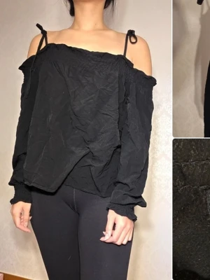 Svart offshoulder topp från Monki - Trendig svart offshoulder topp från Monki med långa ärmar och smala axelband som knyts upptill. Toppen har en lös passform och är tillverkad i ett mjukt tyg som känns skönt mot huden. Perfekt att styla med jeans eller kjol för en cool look.