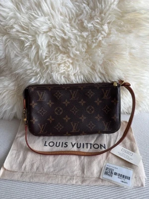Louis Vuitton Monogram Kuvertväska - Snygg kuvertväska från Louis Vuitton i klassiskt brunt monogrammönster. Väskan har en avtagbar axelrem i läder och guldfärgade metalldetaljer. Tillverkad i slitstarkt canvasmaterial med dragkedja upptill. Perfekt för att bära det viktigaste med stil.