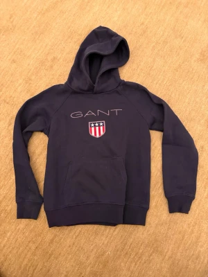 Mörkblå hoodie från GANT - Snygg mörkblå hoodie från GANT med stor huva och känguruficka. Framtill finns GANT-loggan och ett broderat emblem med stjärnor och ränder. Tillverkad i mjuk bomullsblandning, perfekt för en avslappnad och cool stil.