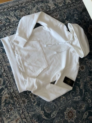 Vit hoodie från Stone Island - Säljer en clean vit hoodie från Stone Island med klassisk huva och känguruficka framtill. Ikoniska Stone Island-märket sitter på ärmen. Tillverkad i mjuk bomull för skön känsla och avslappnad stil. Perfekt för dig som gillar streetwear och vill ha något stilrent.
