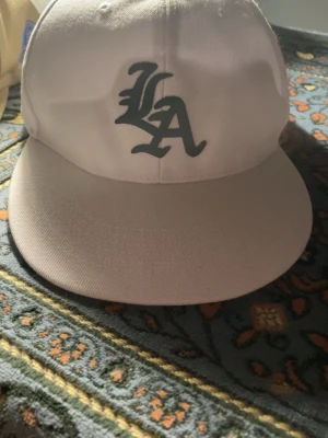 Vit LA keps med broderad logga - Säljer en vit keps med broderad LA-logga i svart framtill och tryckt Los Angeles California-märke på sidan. Klassisk rak skärm och justerbar passform. Tillverkad i bomullstyg, perfekt för dig som gillar streetwear och vill ha en clean look.