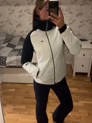 Skidställ från J.Lindeberg i vit och marinblå (storlek S) - Snygg skidjacka och skidbyxor från J.Lindeberg i storlek S. Jackan har gula detaljer på ärmarna och en luva med den gula texten J.Lindberg. Jackan har dragkedja under armarna och fickor för liftkort. Jackan har en fläck på ryggen och är lite färgad av smuts längs dragkedjorna och kanten på ärmarna. Har använt kläderna under några år men nästan bara i fjällen. Skidstället är perfekt för både skidbacken och andra utomhusaktiviteter. Jacka och byxor kan säljas separat också. 