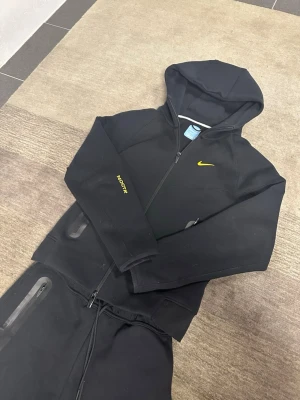 Svart Nike Tech Fleece set med huva - Nike Tech Fleece loungewear set i svart med gul Swoosh-logga på både hoodie och byxa. Hoodien har dragkedja, två fickor med svarta detaljer och huva. Byxorna har dragsko i midjan och dragkedjefickor. Perfekt för chill eller träning.