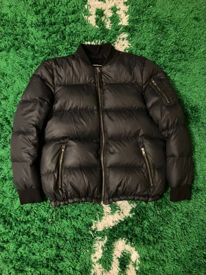 Peak Performance svart dun bomberjacka  - Snygg svart dun bomberjacka från Peak Performance med dragkedja framtill, två fickor med dragkedja och ribbade muddar vid ärmslut och krage. Jackan har en klassisk bomberstil och är perfekt för kalla vinterdagar.