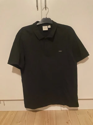 Svart pikétröja från Calvin Klein - Klassisk svart pikétröja från Calvin Klein med liten logotyp på bröstet. Tröjan har korta ärmar, krage och knäppning framtill. Tillverkad i mjukt bomullsmaterial som känns bekvämt mot huden. Perfekt för en clean och stilren look.