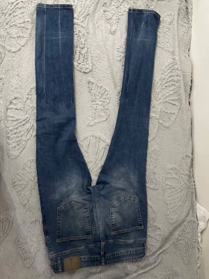 Blå slim fit jeans med slitningar - Säljer ett par blå slim fit jeans med snygga slitningar och tvättade detaljer. Jeansen har klassiska bakfickor och en läderpatch vid linningen. Perfekta för en avslappnad och trendig look.