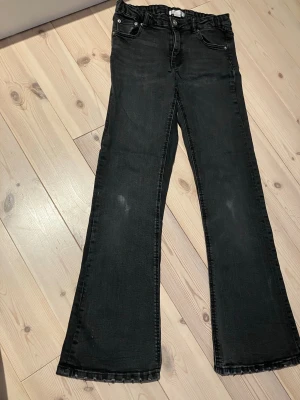 Svarta bootcut jeans med rå kant storlek 158 - Säljer ett par grå/svarta bootcut jeans med rå kant nertill. Jeansen har klassisk femficksmodell, hög midja och utsvängda ben. Materialet är stretchigt denim som sitter snyggt och bekvämt. Perfekta för en trendig och avslappnad look. Två slitningar i tyget se bild inga fläckar. 