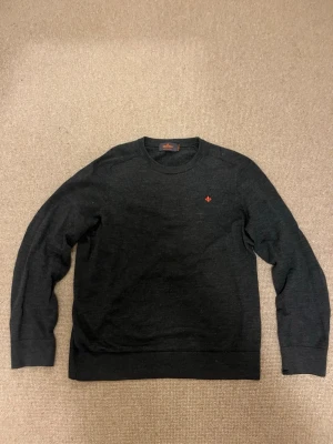 Mörkgrå merinoullströja Morris - Snygg mörkgrå stickad tröja från Morris i 100% merinoull. Tröjan har rund halsringning, ribbade muddar och en liten röd broderad logga på bröstet. Perfekt för dig som gillar stilrena och klassiska plagg. Hör av dig vid prisförslag eller andra funderingar