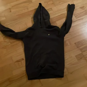 Mörkgrå hoodie från Lyle & Scott - Snygg mörkgrå hoodie från Lyle & Scott med den klassiska guldfärgade loggan broderad på bröstet. Hoodien har ribbade muddar vid ärmslut och nederkant. Perfekt för en avslappnad och stilren look.
